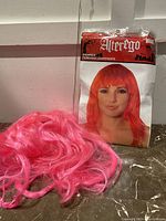 Photo showing the pink wig loose on the floor with the sealed red wig packaging behind it, to demonstrate condition and packaging.