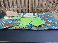 View of the pile of tablecloths on a black surface outside, showing bright floral and pineapple patterned, plain green, and burlap fabrics.