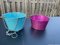 Two tin pails on a black surface outside, one blue with hanging chains and one pink without handles.