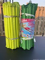 Two folded inflatable float loungers, one lime green and one dark green, secured with straps. Dark green lounger is wrapped in original packaging with product advertising.