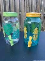 Two beverage containers pictured on black surface with wooden fence background. One clear with green lid and tropical leaf prints, the other teal with yellow lid and pineapple prints. Both have spouts near base.