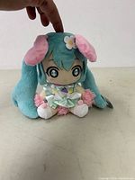 Front view of Hatsune Miku plush doll sitting, showing teal twin tails, pink fluffy wristbands, flower hair clip, and large eye embroidery.