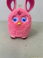 Front view of pink Furby Connect 2016 with eyes open, showing electronic eyelids with blue and purple colors, yellow beak, and large rabbit-like ears with purple interior.
