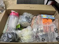 Large cardboard box filled with various miscellaneous kitchen and household items including a pink shaker bottle, citrus juicer, and items wrapped in plastic and bubble wrap.