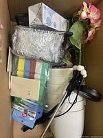 View of box contents showing black metal tool with plastic wrap, seed packets, white plastic bucket with lid, blue tin, colorful straps, pink faux flowers, and other miscellaneous items.