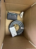 Top-down view of box showing a coiled gray hose with brass fittings, small box, and a metal tool.