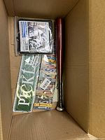 Interior of box showing metal license plate signs, packaged automotive mirror accessory, a red cylindrical item, and a long thin metal rod with a ball end