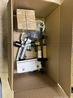 Top view of large box with miscellaneous new and some used household, auto, and electronic parts including metal spray nozzle, boxed automatic drain stop, small boxes and plastic-wrapped items.
