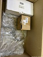 Box with scattered new and some possibly used miscellaneous items including electronics, tools, and household supplies packed with protective plastic.