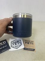 Hand holding navy blue stainless steel YETI promotional cup with embossed logo on the side alongside promotional stickers and registration info cards.