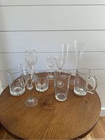 Ten assorted crystal and clear glassware pieces displayed on wooden table, various styles and sizes including stemmed and handled glasses.