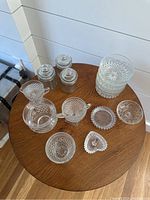 Lot of 14 glass and crystal pieces arranged on a round wooden table showing canisters, creamer, ring dish, coasters, and bowls with cut designs.