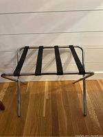 Metal frame luggage rack with black fabric straps placed against a white wall on wooden floor.