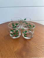 Four short drinking glasses with green holly leaf and red berry hand painted decoration, placed on wooden surface.