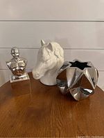 Image showing silver bust sculpture, white ceramic horse head figurine, and silver geometric vase arranged on a wooden surface.
