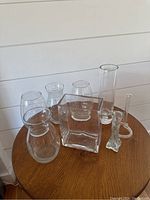 Photo showing 8 clear glass vases of varying shapes and sizes arranged on a wooden table.