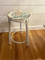 Front angle view showing the full stool with three wicker-wrapped legs and circular wicker footrest ring, distressed white paint with signs of wear.