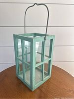 Wooden square candle votive holder with metal handle, shown from an angled view on a round wooden table, background is white shiplap wall.