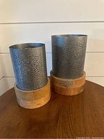 Two perforated metal candle holders with wooden bases standing side by side on a wooden surface, showing front view.