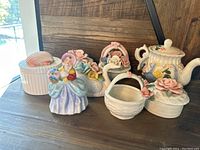 Seven ceramic decorative items including figurines, baskets, teapot, and lidded containers with floral details.