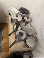 Cookware and kitchen tools laid out on a wooden surface including pots, pans, casserole dishes, strainer, and salad spinner.