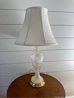 Full view of the lamp showing the clear ribbed crystal body, gold metal base, and white fabric lampshade on a wooden surface.
