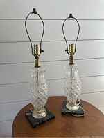 Pair of two clear crystal table lamps with diamond-cut pattern on bodies, gold-tone hardware, and black marble bases shown on wooden table against white shiplap wall.
