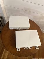 Two closed white metal first aid kits shown on a round wooden table with clasps and wall mounting brackets visible at the back.