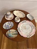 Overview of 8 ceramic pieces including plates, vase, covered cup, and butter dish arranged on round table.
