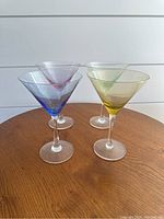Photo showing four multi-colored martini glasses on a wooden surface with a white shiplap wall backdrop. Colors include blue, pink, green, and yellow.