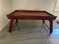 Side angled view of the wooden serving tray showing foldable leg and raised frame with wicker rattan surface