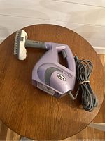 Purple Shark handheld garment steamer with attached fabric brush pad and power cord coiled beside it on wooden table.
