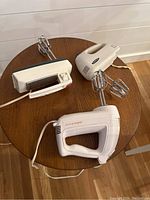 Overhead view showing three electric hand mixers on a round wooden table, all with chrome beaters attached.
