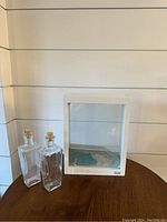 White wooden forever frame with double pane glass and two glass bottles with cork stoppers on wooden tabletop.