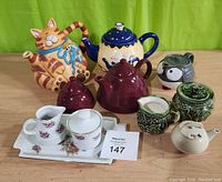 Full lot of seven items on wooden surface with green backdrop showing all teapots and cream sugar sets.