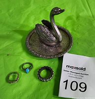 Photo of three rings and a metal swan ring holder on a green background with MaxSold number card.