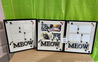 Three black photo frames with white backgrounds featuring paw prints and 'MEOW' text, arranged on a wooden surface in front of a green curtain. Two frames hold photos, one frame is empty with missing foam spacer visible.