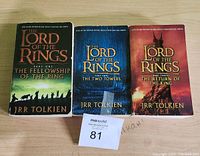 Photo showing three Lord of the Rings softcover books by J.R.R. Tolkien placed side by side on a wooden surface with a MaxSold tag '81' in front.