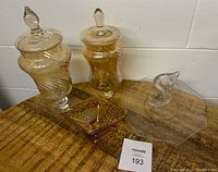 Two clear glass apothecary jars with lids, amber carnival glass bowl, and clear fish-shaped platter arranged on wooden surface.