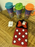 Image showing three reusable Disney-themed plastic cups with handles in blue, green, and orange lids, displayed alongside a Disney kitchen mitt.