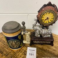 Photo showing the decorative elephant clock and the vintage beer stein on a wooden surface with a lot number tag.