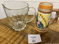 Clear glass pitcher with faceted pattern, handle and pouring spout next to ceramic Florida stein on wooden surface.