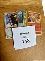 Front view of several Pokémon cards in plastic sleeves showing Moltres and Magearna cards among others, with a MaxSold lot number card in front.