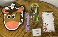 Oven mitt shaped like a Disney animal character, Pixar pen set unopened, purple spoon, and Minnie Mouse notepad visible on wooden table.