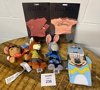 Three Disney nuiMos plush dolls with two sets of doll clothing in original packaging and a pair of Mickey Mouse socks arranged on a wooden table.