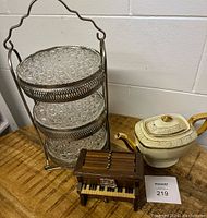 Photo showing the three-tiered glass cake plate stand with metal frame, Hall China teapot with gold accents, and a miniature wooden piano on a wooden surface.