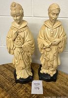 Front view of two vintage cream-colored religious statues on black bases showing detailed robe designs and peaceful expressions.