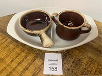 Photo of a white Pyrex divided dish holding two brown McCoy pottery pieces: a handled bowl and a handled cup. The set rests on a wooden surface with a MaxSold lot number card visible.