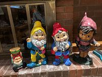 Four ceramic gnome figurines placed on brick hearth arranged left to right: small mushroom-hat gnome, medium yellow-hat gnome, medium red-hat gnome, and larger pink-hat figure.