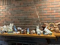 Wide shot of all ceramic figurines arranged on wooden shelf against brick wall, showing various animal and human figures.